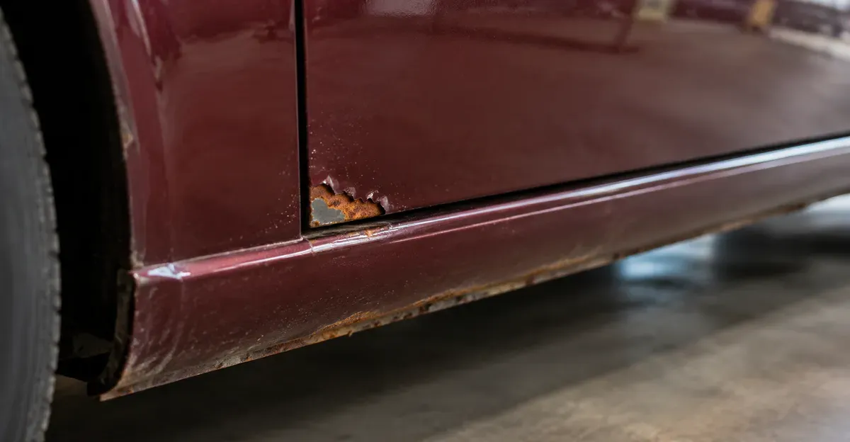 Close-up of surface rust starting at a paint chip on a classic body panel near a door sill, no captions or logos