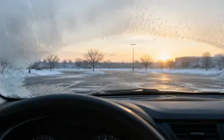 Driver view of a frosted windshield with a partial clear patch over a winter lot at dawn